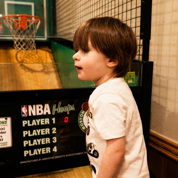 A young boy with brown hair stands in front of an NBA Hoops arcade game.