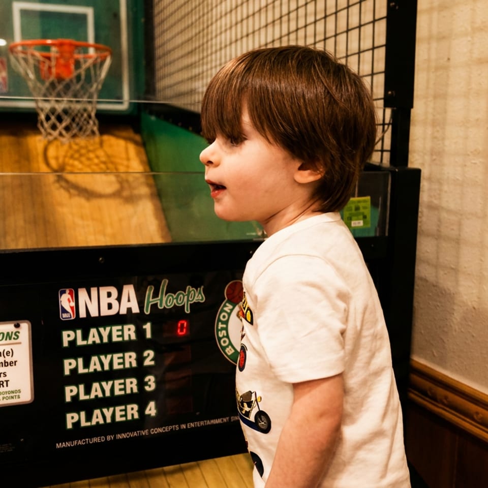 A young boy in a white shirt stands next to an NBA Hoops arcade game, looking intently at the screen.