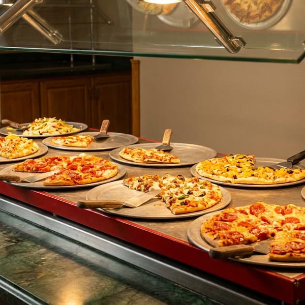 A buffet line with various pizzas on metal trays, some with slices removed, under warming lights.