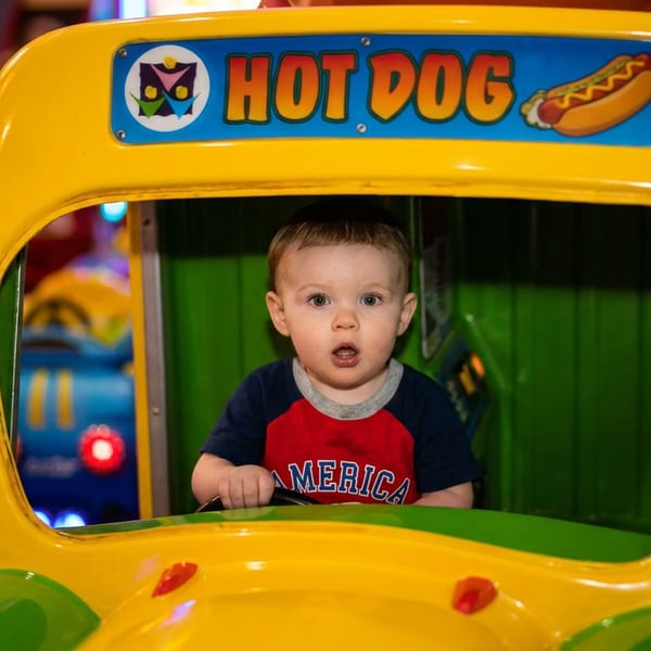 A young boy in a red and blue shirt sits in a yellow hot dog-shaped kiddie ride.