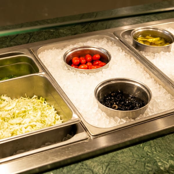 Salad bar with shredded lettuce, spinach, cherry tomatoes, olives, and pickled peppers on ice.