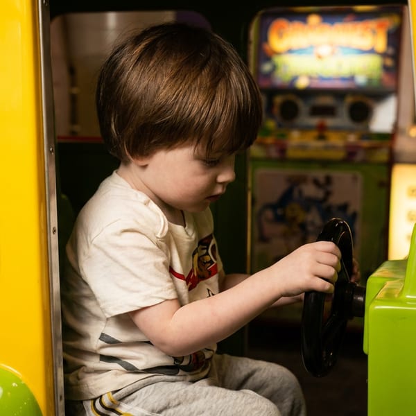 A young boy with brown hair sits in a yellow and green arcade car, holding the steering wheel.