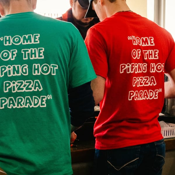 Two people wearing green and red t-shirts that say "Home of the Piping Hot Pizza Parade".