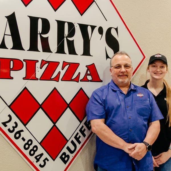 Three smiling Larry's Pizza employees, two women and one man, stand beside a large Larry's Pizza sign.