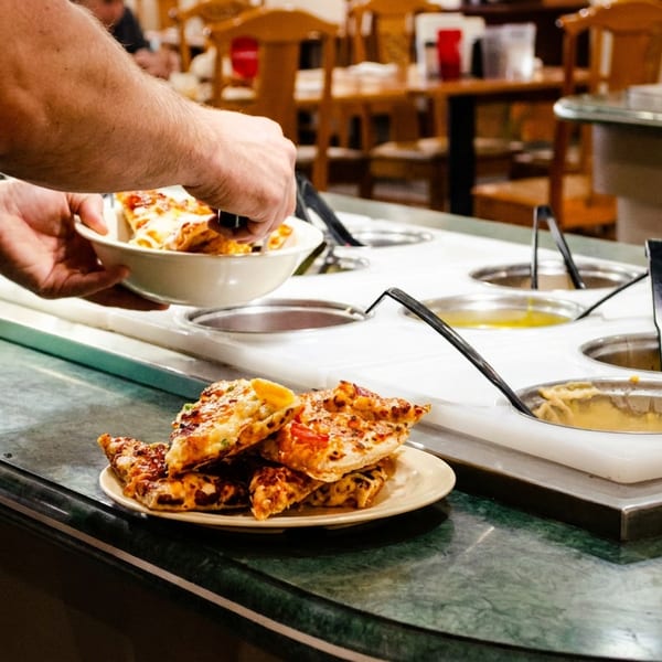 A person serves themselves pizza from a buffet line with various hot food containers.