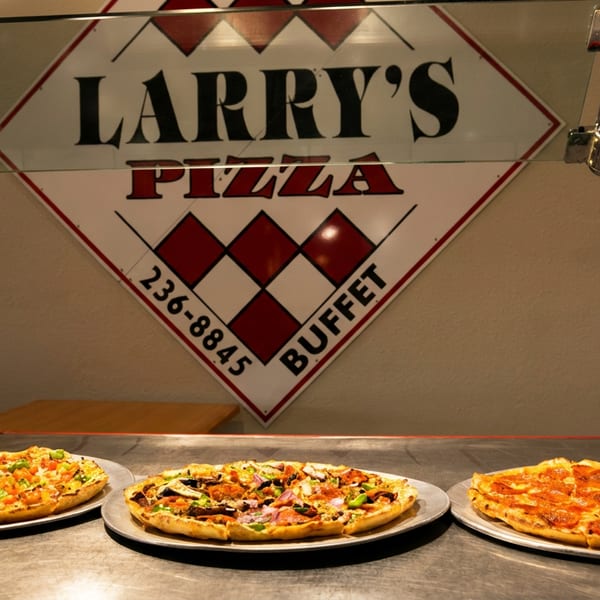 Three pizzas on metal trays sit on a buffet line beneath a "Larry's Pizza Buffet" sign.