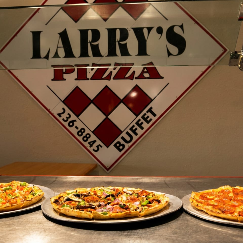 Three pizzas on metal trays sit on a buffet line beneath a "Larry's Pizza Buffet" sign.