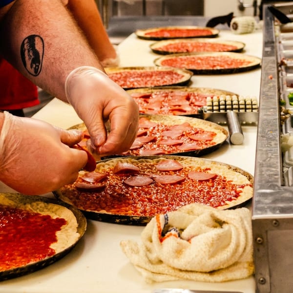 A chef in blue shirt and gloves tops pizzas with pepperoni on a long counter.
