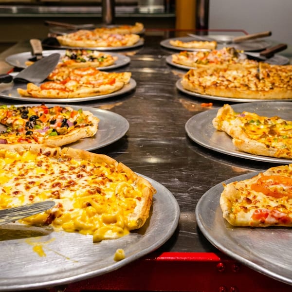 A buffet line of various pizzas on metal trays with serving spatulas.