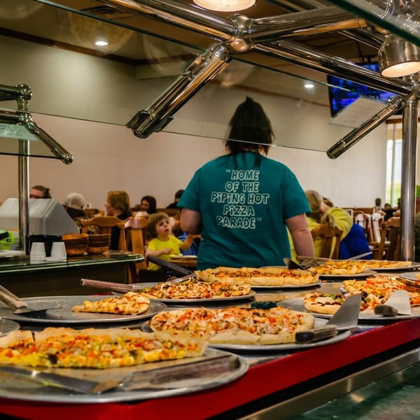 A pizza buffet with various pizzas and a person in a "Piping Hot Pizza Parade" shirt.