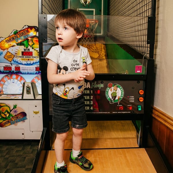 A young boy stands in front of a basketball arcade game, with other arcade games in the background.